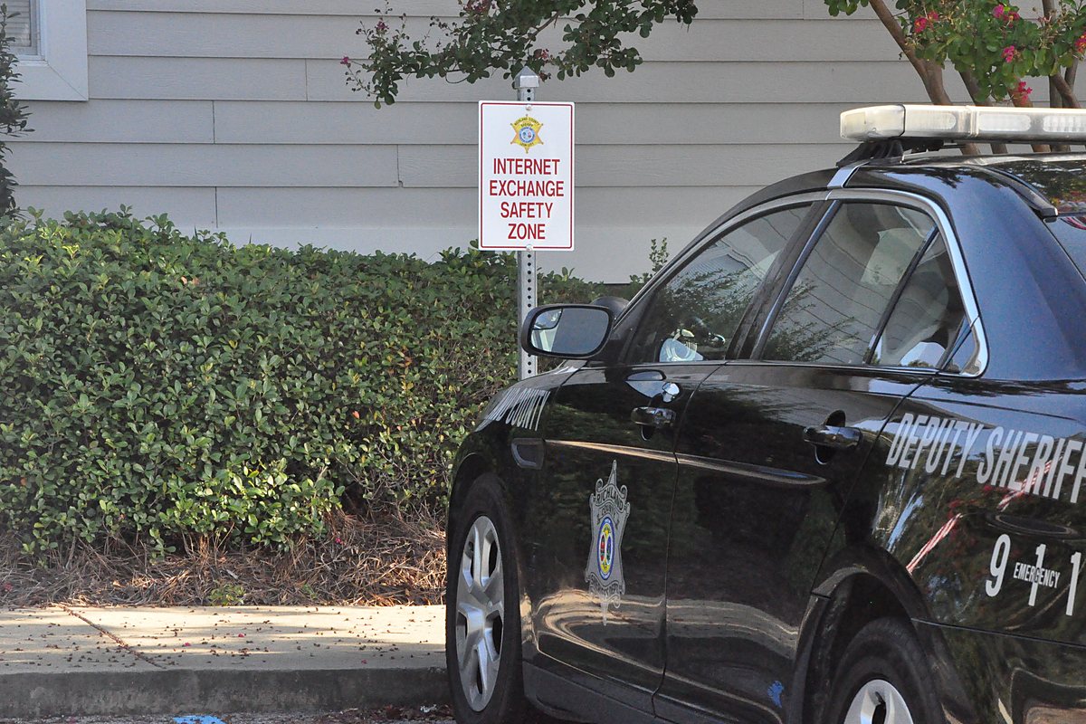An Internet Exchange Safety Zone sign on a post beside a county sheriff's patrol car, outside the sheriff's office