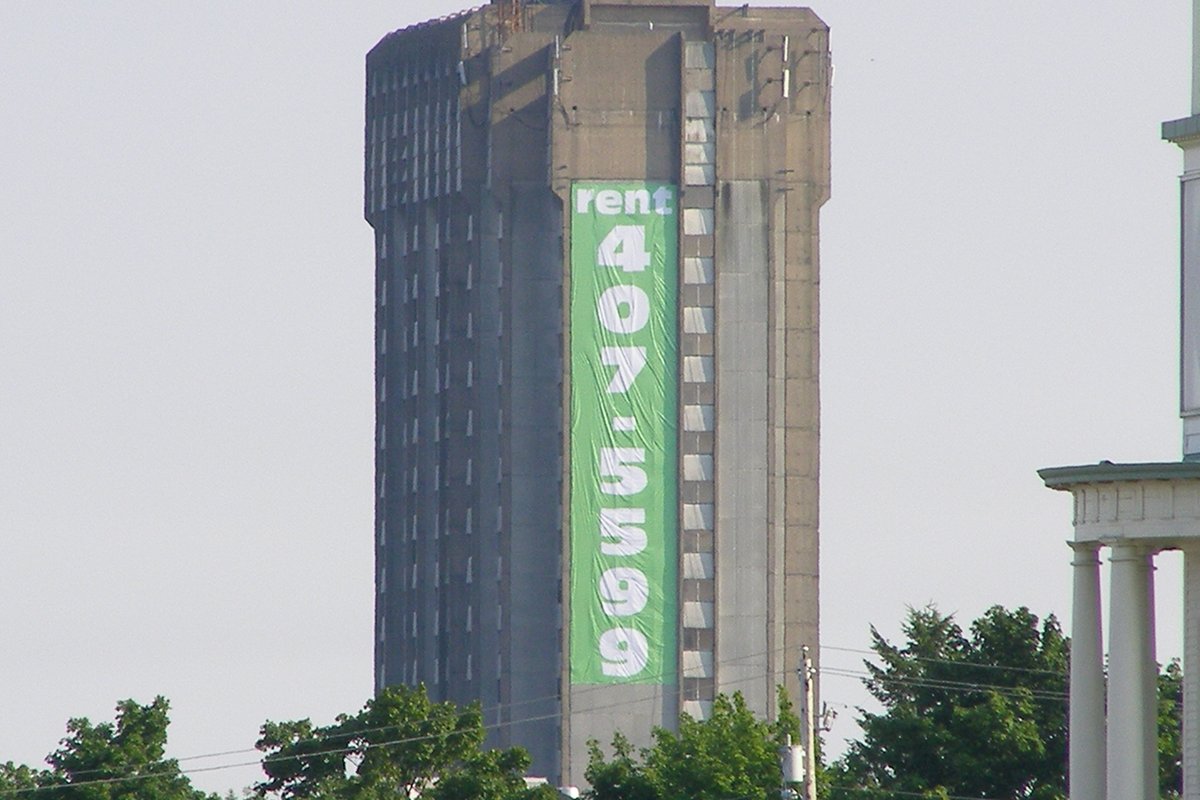 A tall concrete apartment tower with a green banner reading rent and a phone number hung down its side, seen above treetops